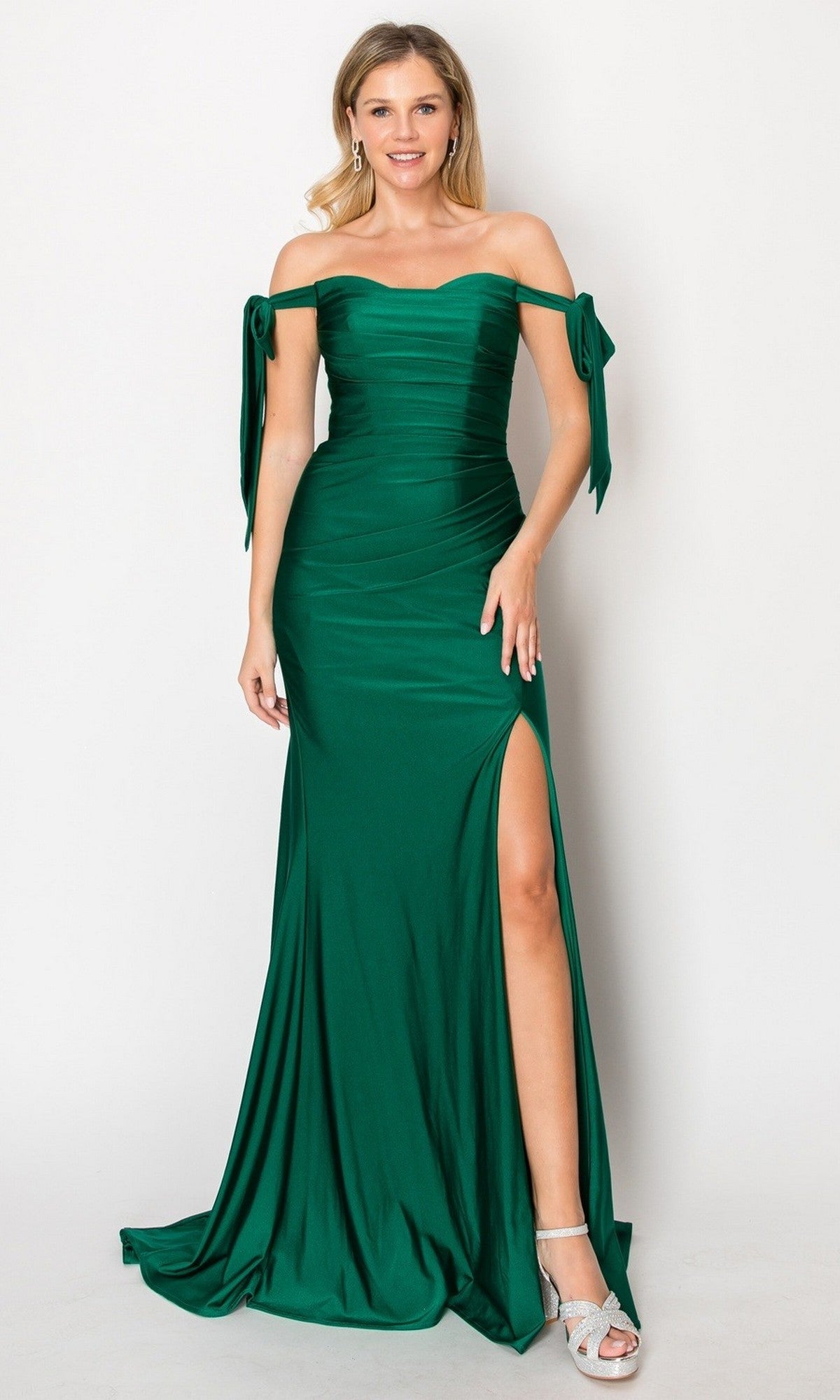 A woman wearing the Long Prom Dress: Chicas DM8010 in green, featuring an off-the-shoulder design with a ruched bodice, bow details on the sleeves, a thigh-high slit, and floor-length silhouette, stands against a plain background.